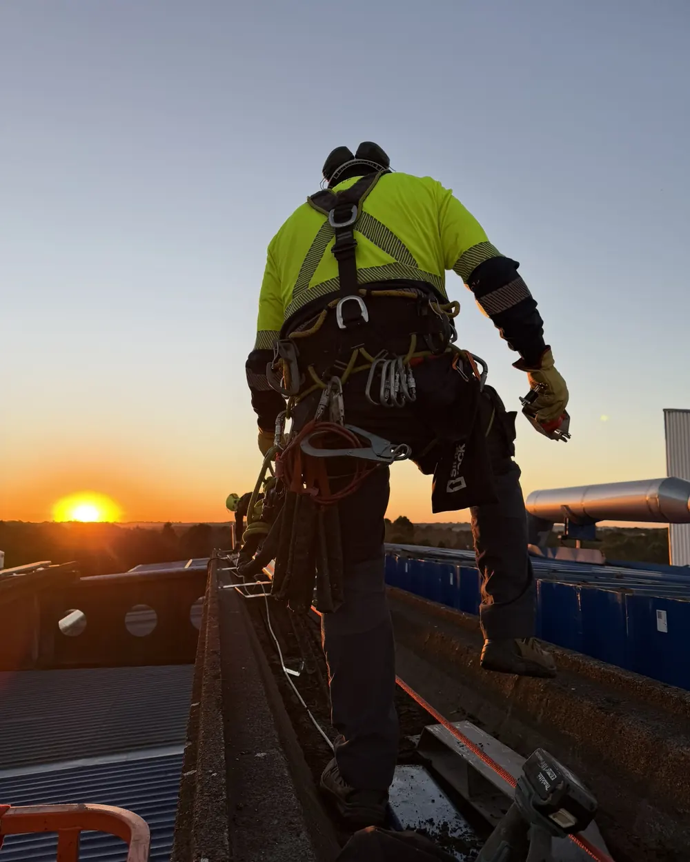 Técnico de VERTEC con arnés y equipo de acceso por cuerdas sobre la cubierta de una planta industrial al atardecer